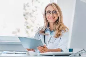 smiling female doctor sitting her desk holding digital tablet