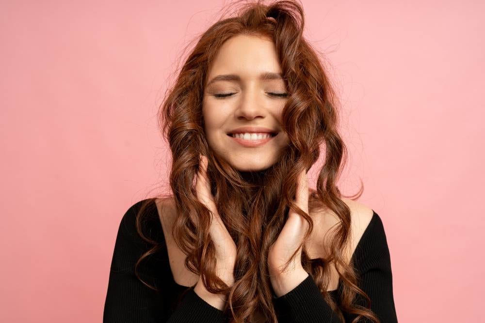 beautiful red head woman with close eyes posing pink wall wavy hairs perfect smile